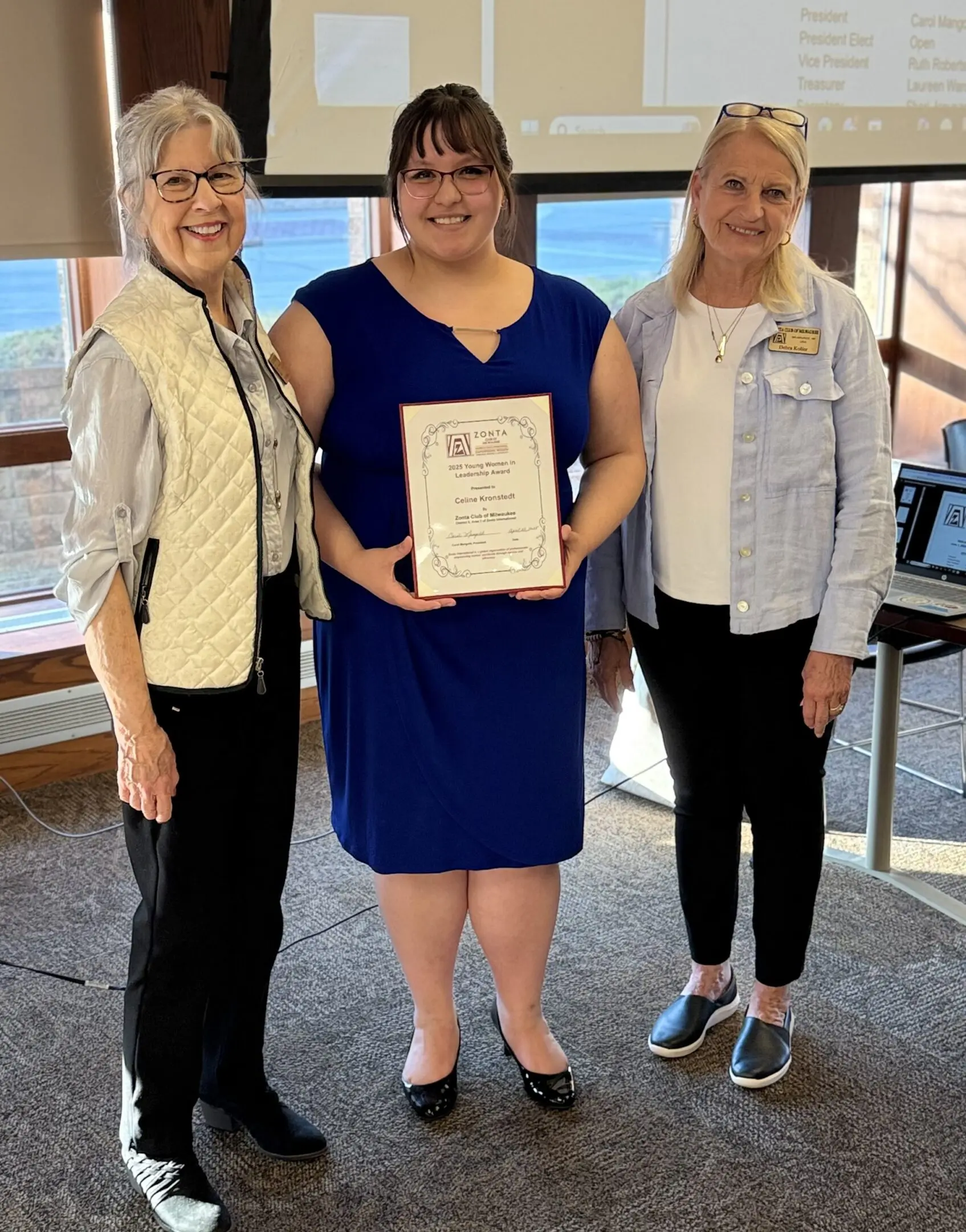 Three women standing indoors, one holding a certificate.